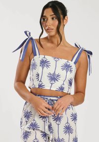 Top corto con estampado de palmeras en azul sobre un fondo blanco. Presenta tirantes de lazo a rayas en azul y blanco. Textura suave.