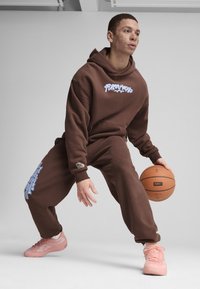 Hnedá mikina s bielym logom, zodpovedajúce hnedé tepláky a ružové športové topánky. Model drží basketbal v dynamickej póze.