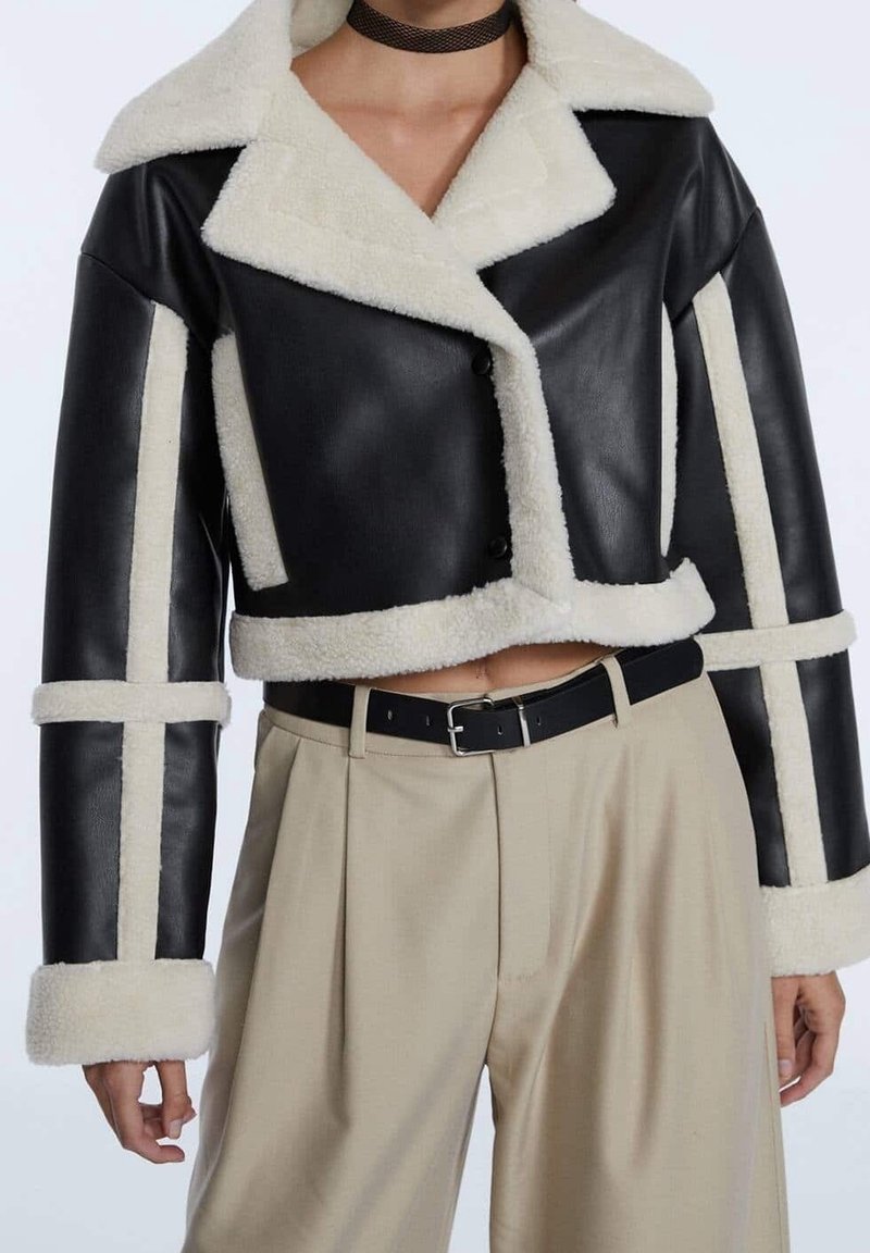 Chaqueta de cuero negra corta con cuello y ribete de piel de oveja blanca. Presenta un cierre de botón y detalles estampados en las mangas, combinada con pantalones beige.