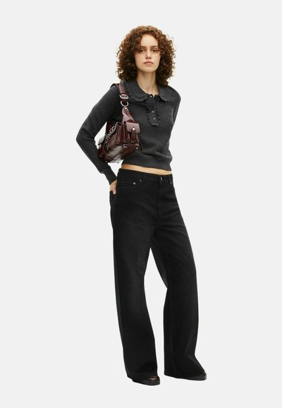 Vrouw met krullend haar die een donkergrijze trui met een gegolfde kraag en brede zwarte broek draagt, terwijl ze een donkerbruin leren schoudertas draagt.