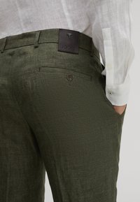 Olijfgroene broek van gestructureerde stof, een achterzak met een knoop en een zwart leren merklabel met een embleem.
