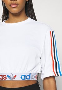 Bílý zkrácený tričko s krátkými rukávy. Má barevné pruhy v červené, černé a modré barvě na rukávech. Elastický pas s logem "adidas".