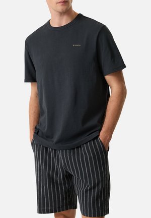 Homme portant un t-shirt noir à manches courtes avec un petit logo et un short noir avec des rayures verticales blanches, debout les mains dans les poches.