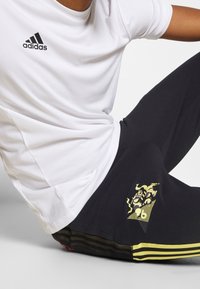 Vit herr t-shirt med fukttransporterande egenskaper och svart adidas-logotyp, i kombination med svarta träningsbyxor med gula ränder och dekorativa detaljer.