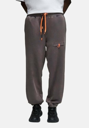 Personne portant un pantalon de jogging gris foncé avec des cordons orange et une poche boutonnée, associée à des baskets noires et un haut blanc.