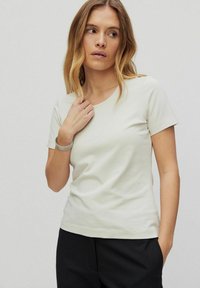 T-shirt i blød beige med rund hals og korte ærmer, der har en glat tekstur og et enkelt design. Sammen med sorte bukser.