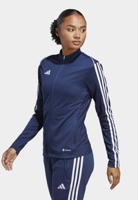 Marineblaue Sportjacke mit weißen Streifen, Reißverschlussfront und strukturierten Seitenpanels. Mit einem Logo auf der Brust und an den Ärmelbündchen.