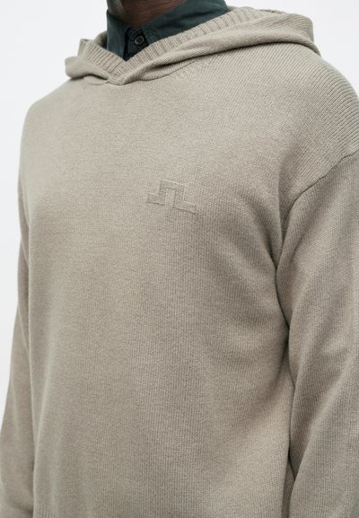 Ανοιχτό γκρι πλεκτή κουκούλα με V-λαιμό, ribbed άκρη και έναν διακριτικό λογότυπο στο στήθος. Μαλακή υφή και χαλαρός σχεδιασμός.