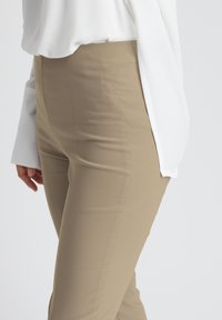 Beige, fitted trousers with a smooth texture and side pockets, paired with a loose white blouse. The fabric appears stretchy and comfortable.