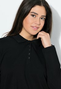 Černá polokošile s dlouhým rukávem a límečkem, vybavená knoflíkovou légou a pružnými manžetami. Vyrobena z měkké, texturované látky.