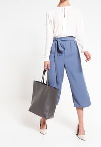 Blouse blanche à manches longues avec ouverture en forme de goutte dans le dos, associée à un pantalon bleu taille haute et court orné d'un nœud. Sac cabas en cuir gris, talons nus.