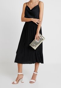 Kobieta w czarnej plisowanej sukience midi na cienkich ramiączkach, trzymająca metaliczną torebkę kopertową i nosząca białe sandały na obcasie z odkrytymi kostkami.