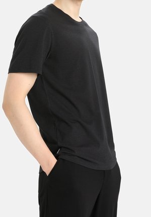 Mann trägt ein schlichtes schwarzes Kurzarm-T-Shirt und schwarze Hose, eine Hand in der Tasche, steht vor hellem Hintergrund.