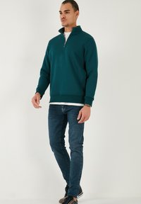 Golfino teal con mezza zip, polsini e fondo a coste. Indossato con jeans blu scuro e scarpe nere con strisce bianche.