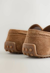 Chaussures en daim brun avec un dos bas, dotées de semelles en caoutchouc texturé et de points de traction ronds, montrant un détail de couture le long des coutures.