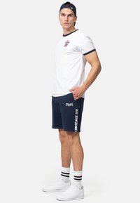 Bijela pamučna majica s mornarsko plavim detaljima, uparena s mornarsko plavim šortsem s logotipom. Bijele tenisice i prugaste sportske čarape upotpunjuju izgled.