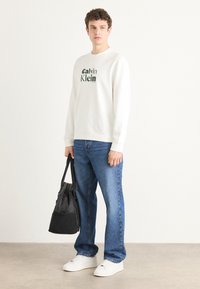 Biely sveter s zeleným nápisom "Calvin Klein", modré džínsy, biele tenisky a čierna taška s obdĺžnikovým tvarom. Jednoduché pozadie.