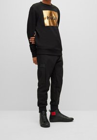 Sweat-shirt noir avec un grand logo doré, pantalon cargo noir avec poches latérales, et baskets montantes noires avec des accents rouges.