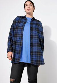 Camicia in flanella a quadri blu e nera, con bottoni e un fit rilassato, indossata sopra una t-shirt blu e abbinata a jeans neri strappati.