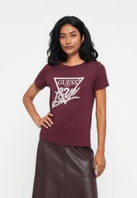 Bordeaux katoenen t-shirt met een ronde halslijn, korte mouwen en een driehoekig "GUESS" logo in crèmekleurige en zilveren versiering.