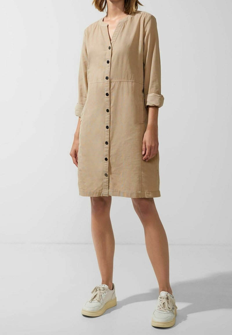 Robe beige à manches trois quarts avec un col en V, dotée de poches et de boutons noirs. Portée avec des baskets beiges.