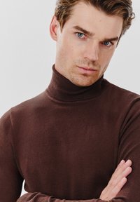Brązowy sweter golfowy wykonany z miękkiego, elastycznego materiału, dopasowany krój, z prążkowanym kołnierzem i gładką fakturą. Ręce skrzyżowane na piersiach.