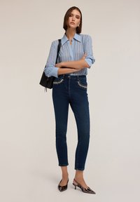 Camisa de botones azul claro con rayas verticales, jeans de mezclilla oscura con acentos de cristal en los bolsillos y zapatos negros de tacón medio brillantes.