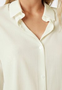Ecru koszula zapinana na guziki, wykonana z teksturowanej, lekkiej tkaniny z delikatnym wzorem zmarszczek i kołnierzykiem w szpic.