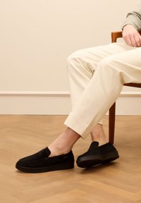 Black suede slip-on shoes with a platform sole, featuring a minimal design and rounded toe, paired with cream-colored trousers.