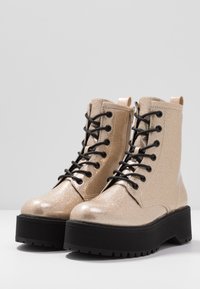 Gouden glitter combatlaarzen met een dikke zwarte plateauzool, voorzien van zwarte veters en een ritssluiting aan de zijkant voor gemakkelijk dragen.