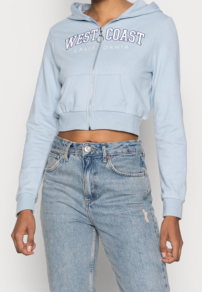 Personne portant un sweat à capuche court bleu clair zippé avec le texte « West Coast California » et un jean bleu clair taille haute légèrement usé.