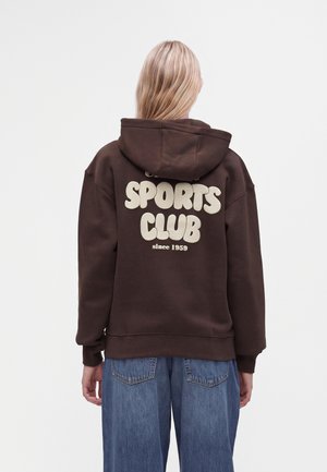 Personne blonde portant un sweat à capuche marron foncé avec le texte "Sports Club since 1959" dans le dos et un jean bleu large, vue de dos.