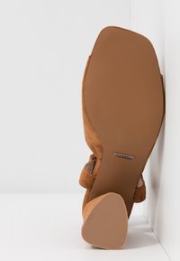 Chaussure à talon haut en daim marron reposant sur sa semelle, montrant un talon texturé et une semelle lisse avec une petite étiquette de marque rectangulaire.