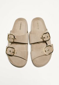 Beige slide sandaler i mocka med två breda remmar, var och en med en stor guldbuckle. Texturerad fotbädd och minimalistisk design.