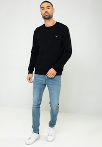 Zwarte sweatshirt met lange mouwen, ronde hals en een klein oranje logo op de borst, gecombineerd met lichtblauwe jeans en witte sneakers.