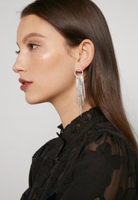 Orecchini pendenti d'argento con base circolare, caratterizzati da molte frange decorate con piccoli strass. Modella che indossa un blusa nera di tessuto texturizzato.