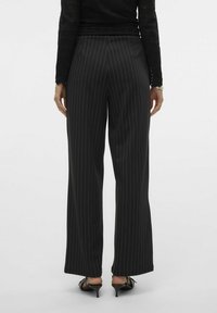 Pantalons noirs taille haute avec des rayures vertes verticales, coupe évasée, tissu lisse et léger brillant, complétés par des talons noirs élégants.