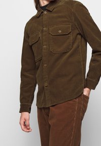 Camisa de pana marrón con collar abotonado, dos bolsillos en el pecho y textura acanalada. Combinada con pantalones de pana marrón a juego.