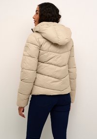 Béžová puff jacket s odnímateľnou kapucňou, s prešívanými vzormi a elastickými manžetami, v kombinácii s tmavými džínsami.
