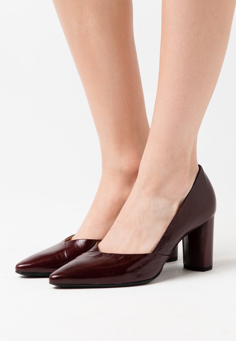 Chaussures à talons hauts bordeaux avec un bout pointu et un talon carré. Le matériau a une finition brillante avec une texture lisse.