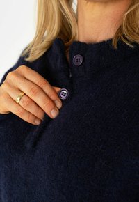 Maglione lavorato a maglia blu navy con scollo rotondo, caratterizzato da due bottoni viola all'apertura e un tessuto morbido e strutturato. Mano che sistema il bottone.