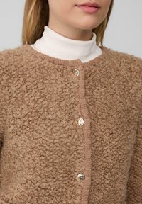 Close-up van een persoon die een beige gestructureerde wollen cardigan draagt met drie glanzende gouden knopen over een witte coltrui.