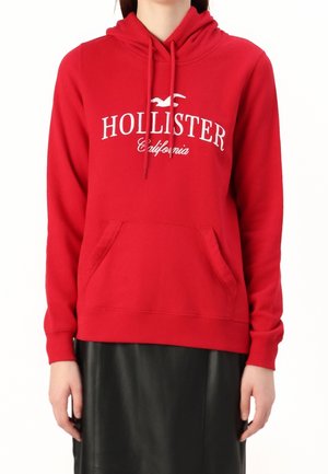 Osoba ubrana w czerwoną bluzę z kapturem Hollister California z przednią kieszenią i czarną spódnicę, przedstawiona od szyi do bioder na białym tle.