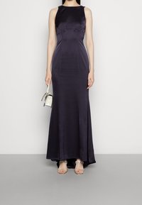 Marinblå satängklänning utan ärmar med en figurnära överdel och utsvängd kjol. Modellen håller en liten vit handväska och bär utsmyckade genomskinliga sandaler.