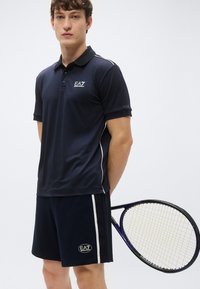 Námornícka polokošeľa s golierom a bielymi akcentmi, doplnená o zladené šortky. Model drží tenisovú raketu. Obe veci nesú logá EA7.