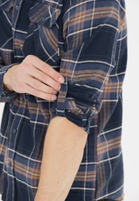 Tummansininen ja ruskea ruudullinen flanellipaita, jonka hihat on käännetty, napitettu rannetuki ja rintatasku. Pehmeä rakenne, rento muotoilu.