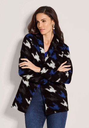 Vrouw met lang donker haar draagt een zwart-blauw-wit patroonjack over een blauw topje en jeans, staat met gekruiste armen en kijkt naar rechts.