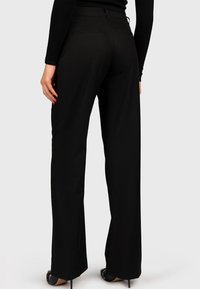 Pantaloni neri larghi realizzati in un tessuto leggero e morbido. Presentano una vita media e pratiche tasche posteriori. Abbinati a tacchi alti neri.