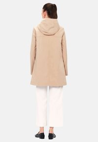 Cappotto beige con cappuccio e vestibilità rilassata, caratterizzato da un taglio dritto e senza motivo visibile. Abbinato a pantaloni bianchi a gamba larga e scarpe nere senza lacci.