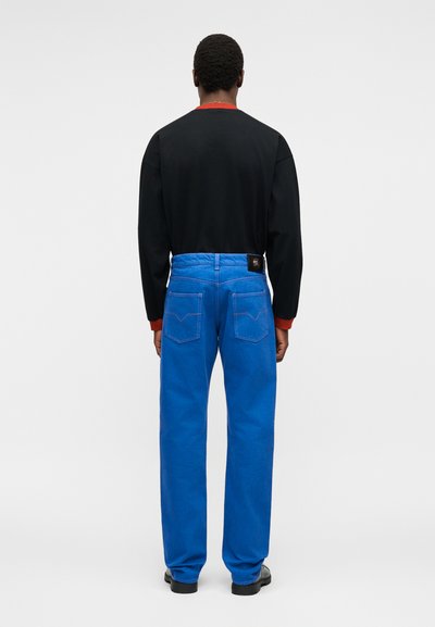 Άτομο που φοράει φωτεινό μπλε τζιν με ψηλή μέση και ίσια ίσια πόδια και μαύρο μακρυμάνικο πουκάμισο με κόκκινες μανσέτες, στέκεται με την πλάτη προς τα εμπρός σε απλό φόντο.